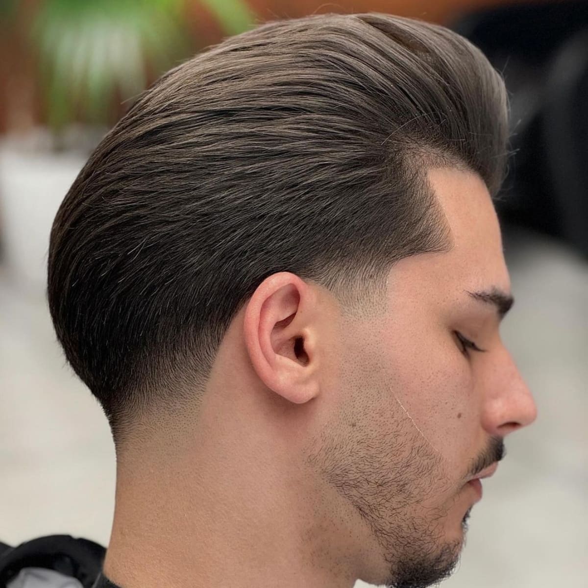 Side profile of a man with a perfectly styled tapered slick back haircut, featuring a sleek, voluminous top and a clean, gradual fade on the sides. His well-groomed beard complements the sharp, polished look, showcasing the seamless blend of hair and facial hair styling.