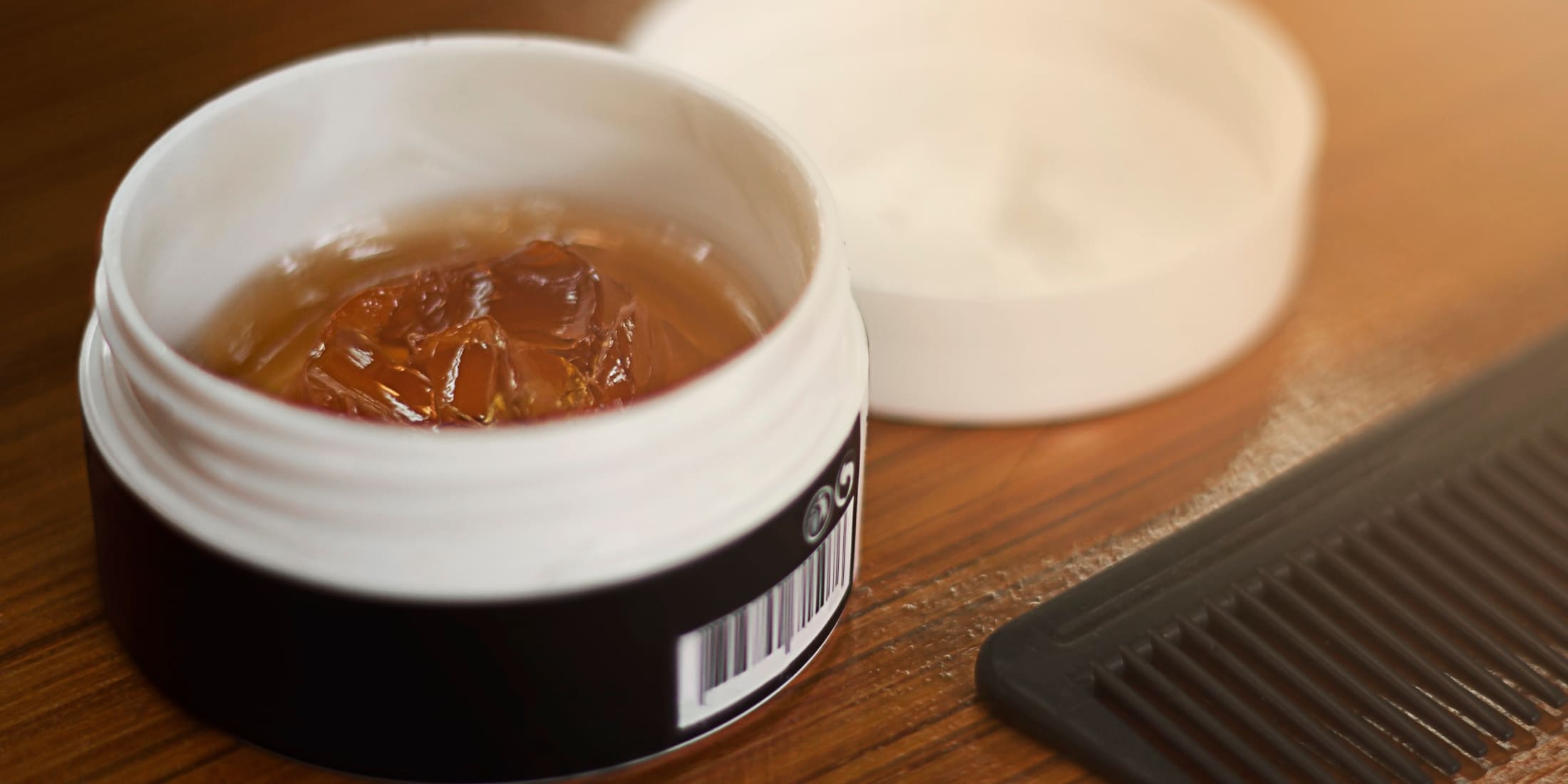 Open container of amber-colored pomade with a white lid and a black comb beside it on a wooden surface, illustrating hair styling product choices and the difference between pomade and gel.