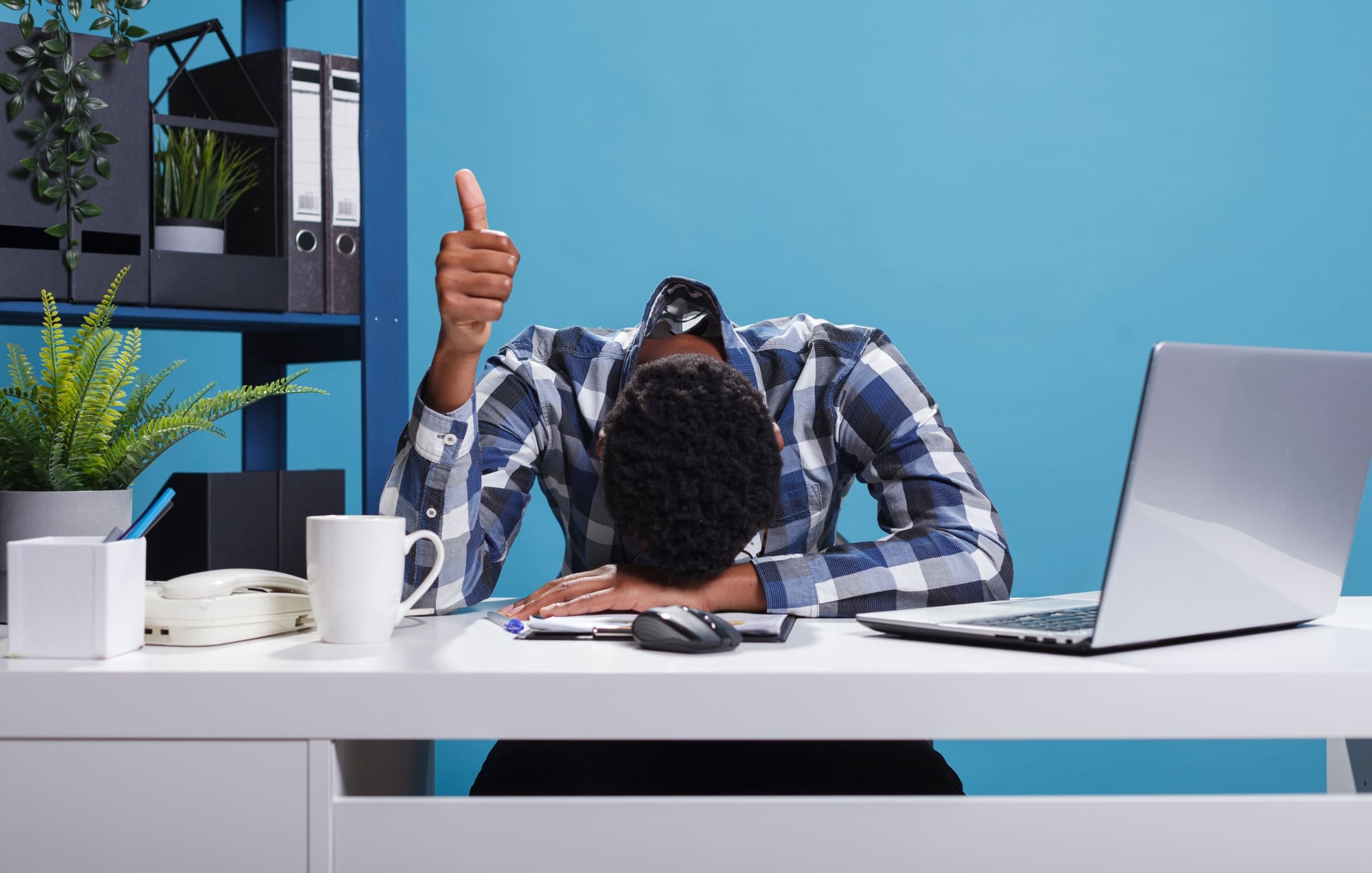 Overworked dad slumped over his desk with a thumbs-up, showing signs of burnout while trying to stay positive—highlighting the importance of self-care for fathers balancing work and family life.