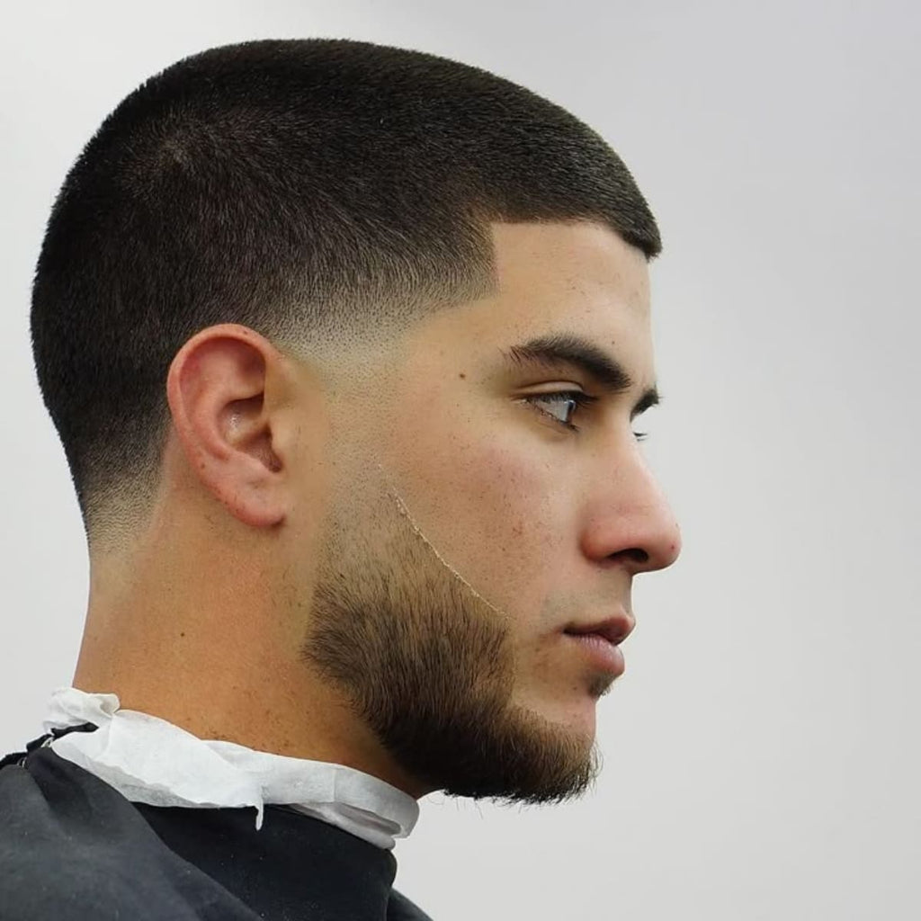 Side profile of a man with a buzz cut low taper haircut. The top is evenly cut short, and the sides feature a smooth low taper fade that starts just above the ears, blending seamlessly into the skin. The style highlights a sharp, clean beard line that complements the haircut for a polished, well-groomed look.