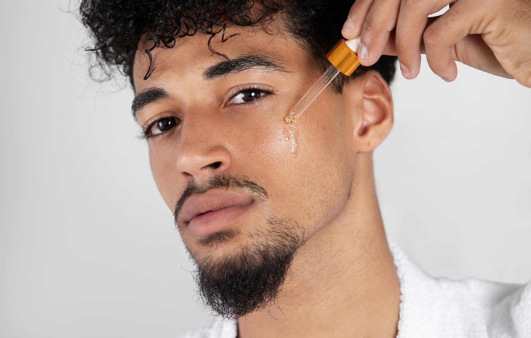 A man applies serum to his face using a dropper, focusing on hydrating and nourishing his skin as part of his men's skincare routine. The serum glistens on his cheek, promoting healthy, glowing skin.