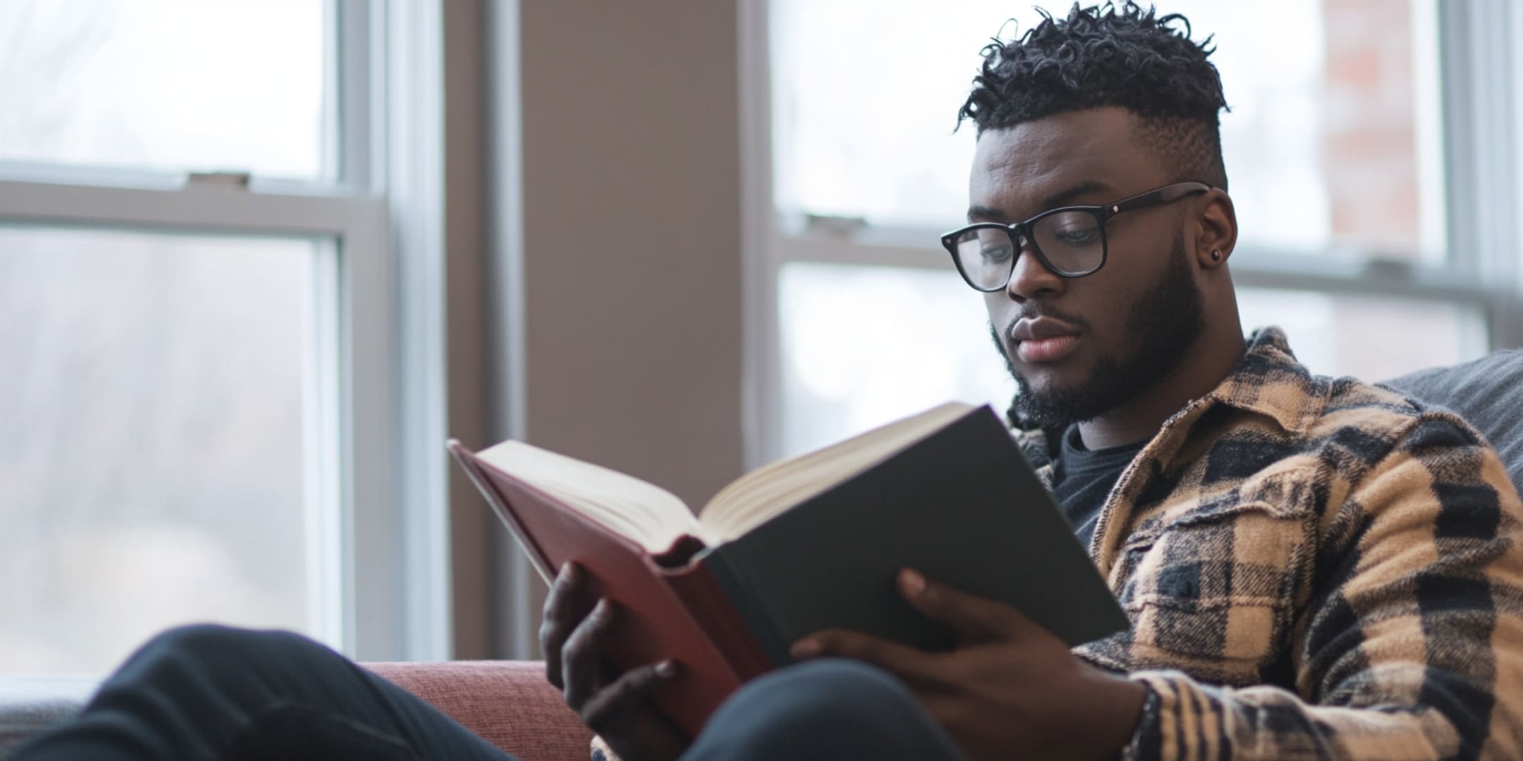 A man wearing glasses and a plaid shirt sits comfortably by a window, deeply focused on reading a large book. This scene represents self-education, one of the habits of successful men, emphasizing the importance of continuous learning and personal growth.