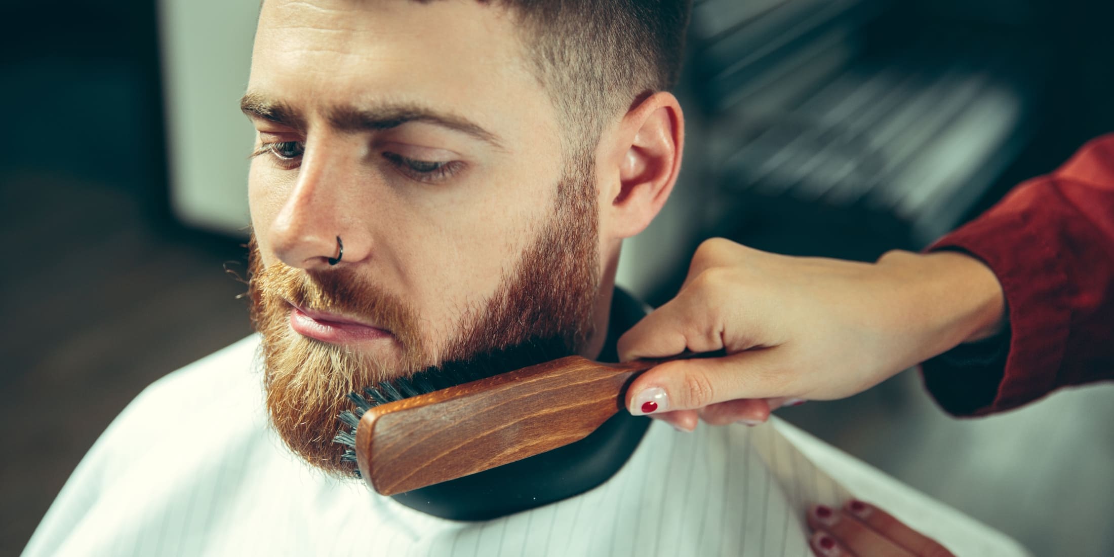 Man getting his beard brushed at a barber shop with a wooden beard brush, highlighting the benefits of brushing your beard. Brushing promotes healthy growth, distributes natural oils, reduces tangles, and enhances beard appearance.