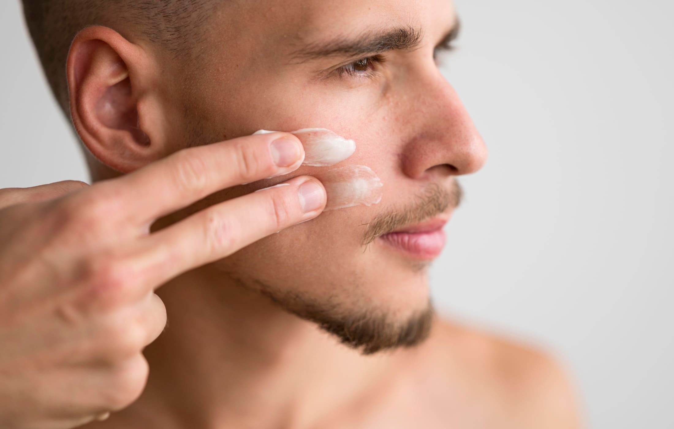 Side profile of a man applying moisturizer to his face, gently smoothing the product into his skin with his fingers. His focused expression highlights the importance of hydration in his skincare routine for a healthy complexion.