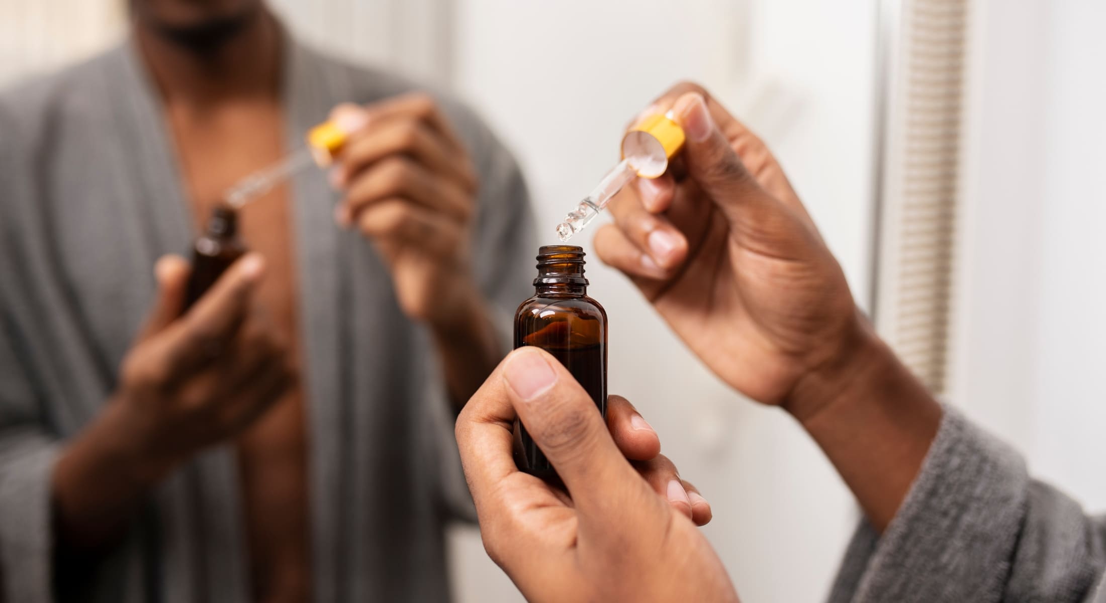 A man in a robe preparing to apply beard oil, holding a dropper and bottle in front of a mirror as part of his grooming routine.