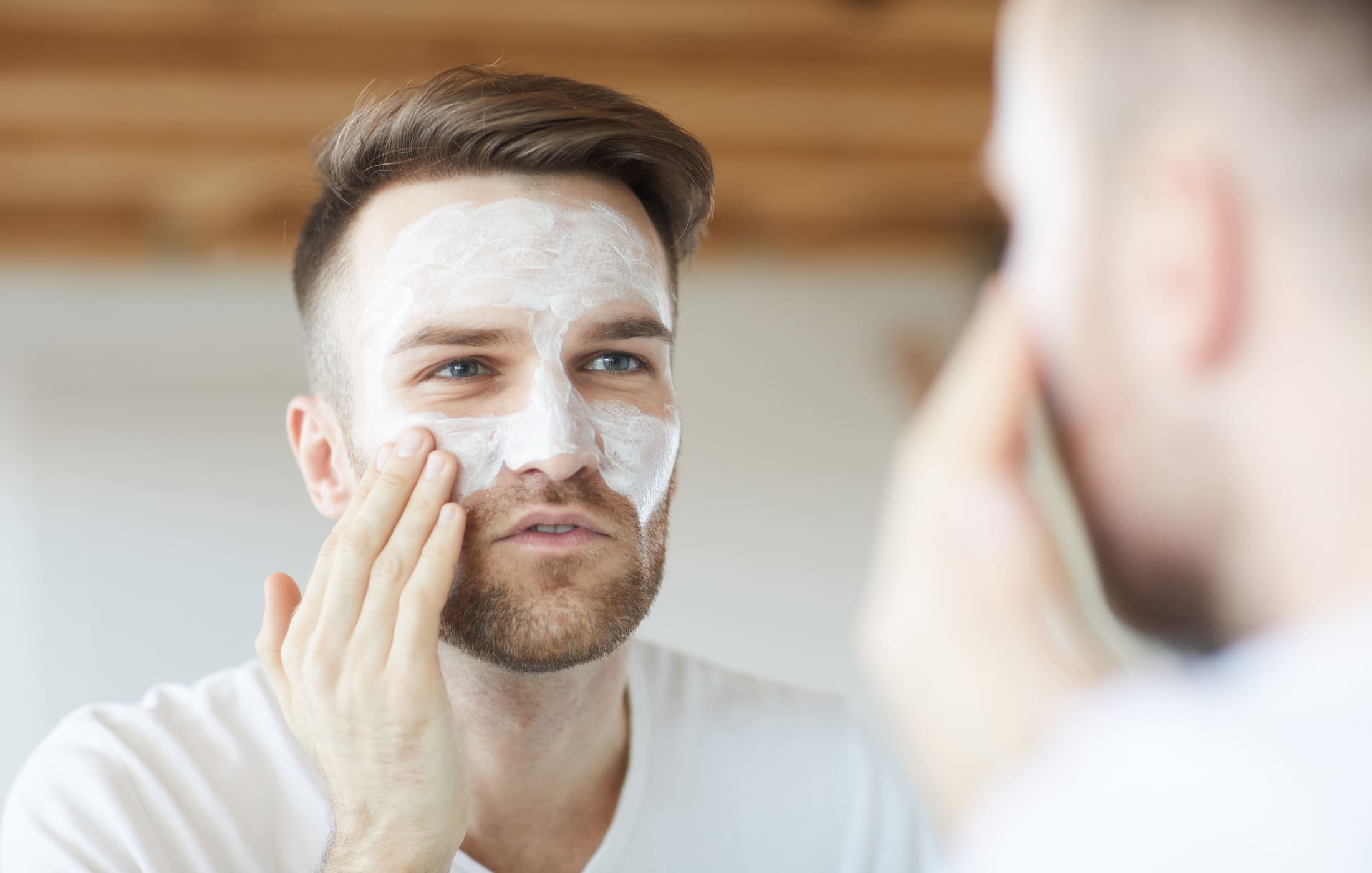 A man applies a white face mask while looking into the mirror, focusing on his routine.