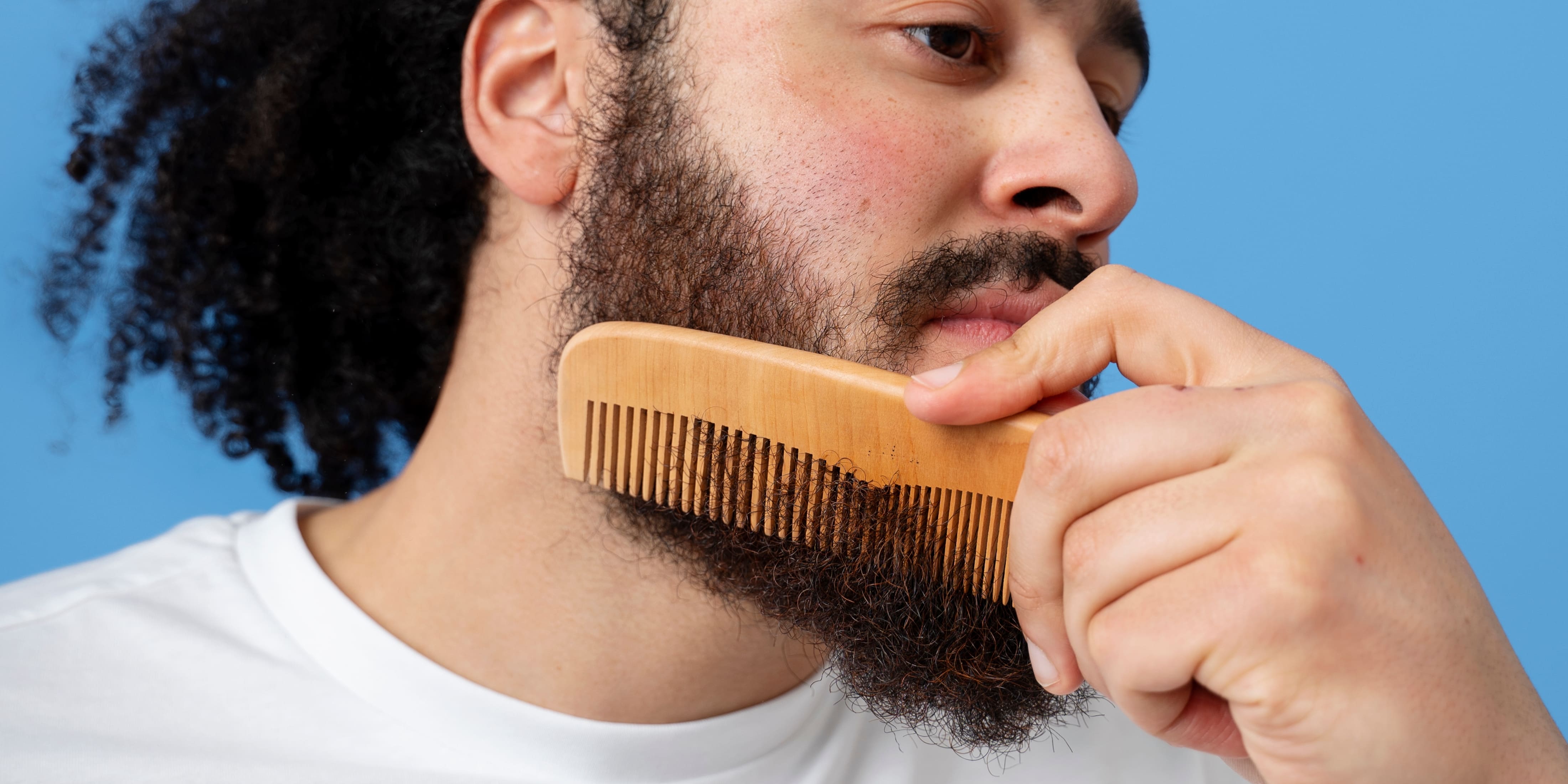 Man using a wooden beard comb to groom his thick beard against a blue background. Ideal for beard care and grooming routines. Beard comb vs beard brush comparison.