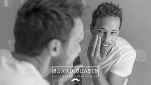 A smiling man applying facial cleanser as part of his men's skincare routine, looking at his reflection in the bathroom mirror.