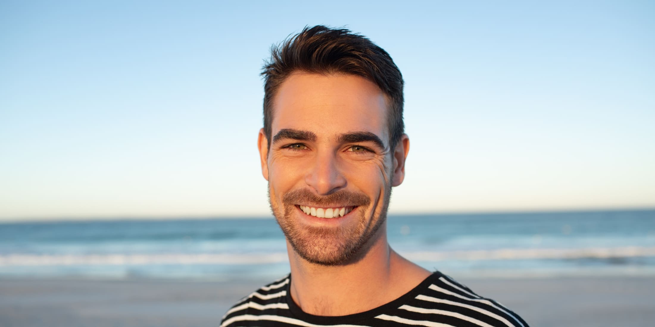 A man with a well-groomed 5mm beard, smiling brightly at the camera. He is wearing a black and white striped shirt and standing on a beach with the ocean and sky in the background, giving the image a serene and relaxed feel.