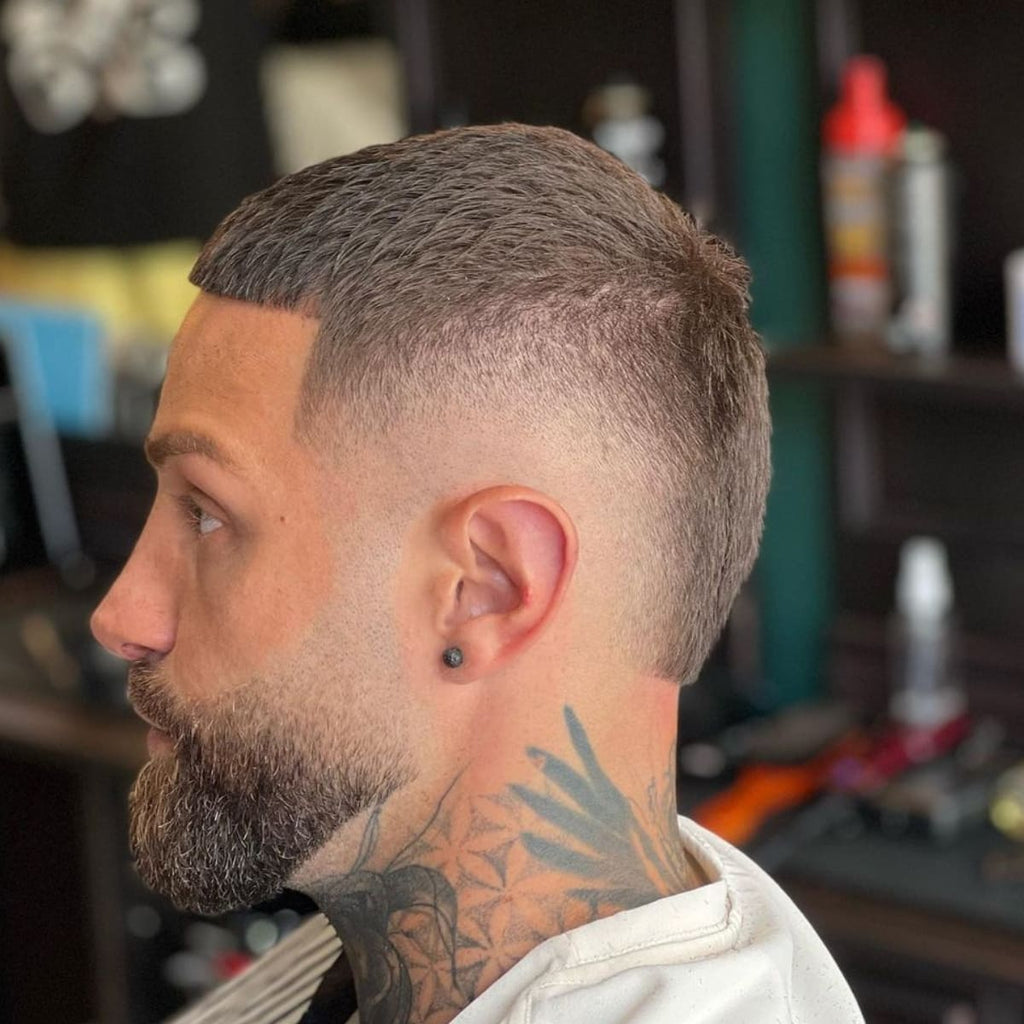 Side profile of a man with a classic burst fade buzz cut, featuring a smooth fade around the ears and a neatly trimmed, short top. The style is paired with a well-groomed beard and visible neck tattoos, giving a sharp and stylish look.