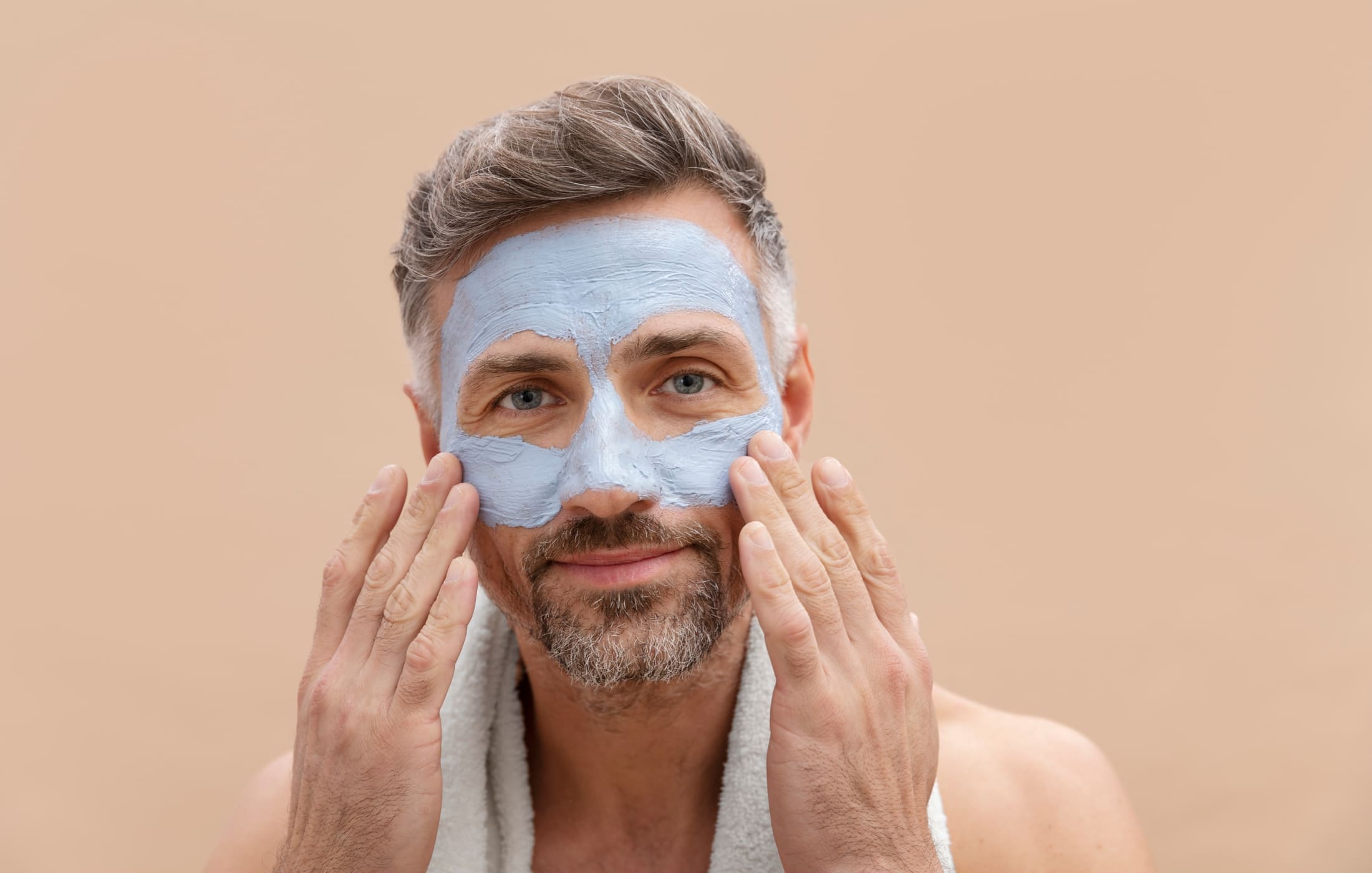 A man with salt-and-pepper hair applies a blue face mask, focusing on his skincare routine.