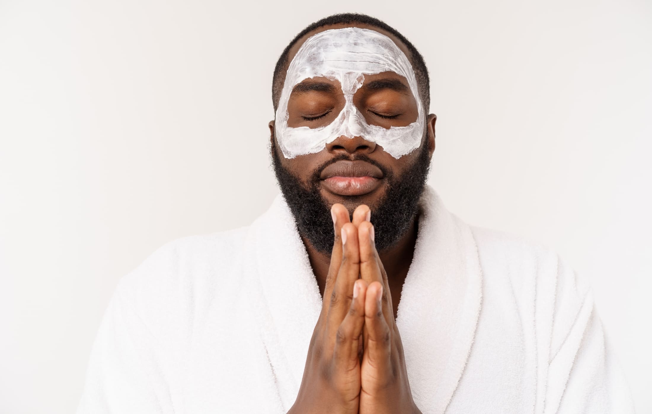 A man peacefully applies a white face mask, with closed eyes and hands clasped in a relaxed, meditative pose.