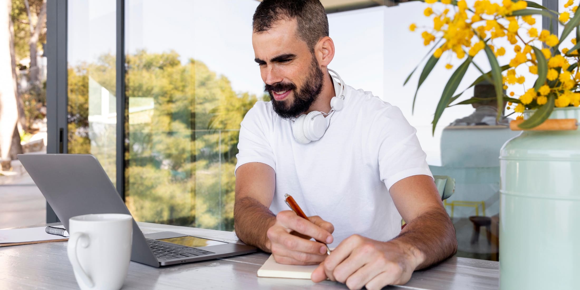 A man working from home, sitting at a desk with a laptop and notebook, jotting down notes while wearing headphones around his neck.