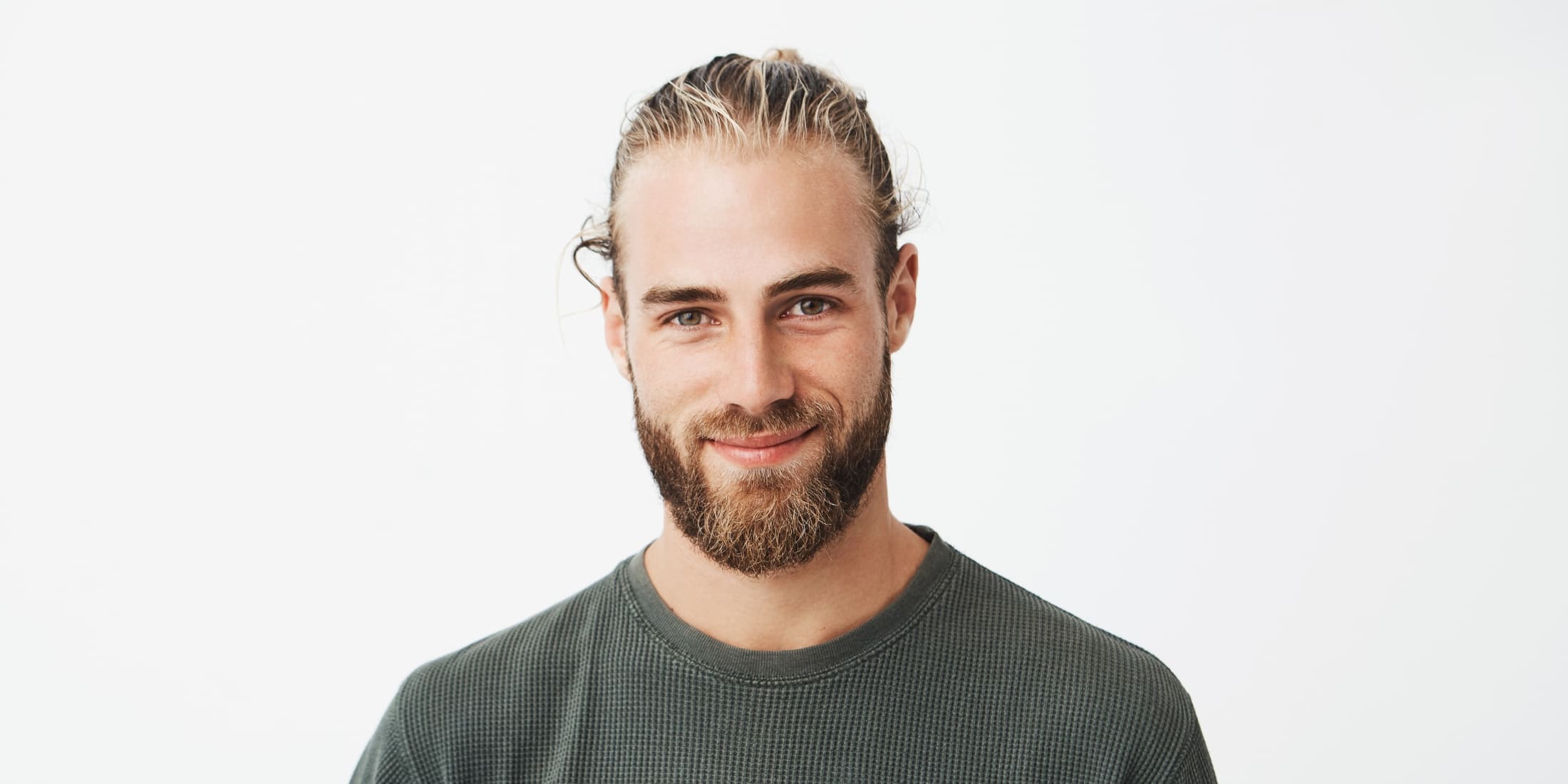 Smiling young man with a well-groomed, mature beard, and long hair tied back, showcasing a confident and natural look. His beard is full and healthy, representing the results of consistent beard care, ideal for highlighting the ongoing benefits of using beard oil to maintain softness, prevent dryness, and promote overall beard health.