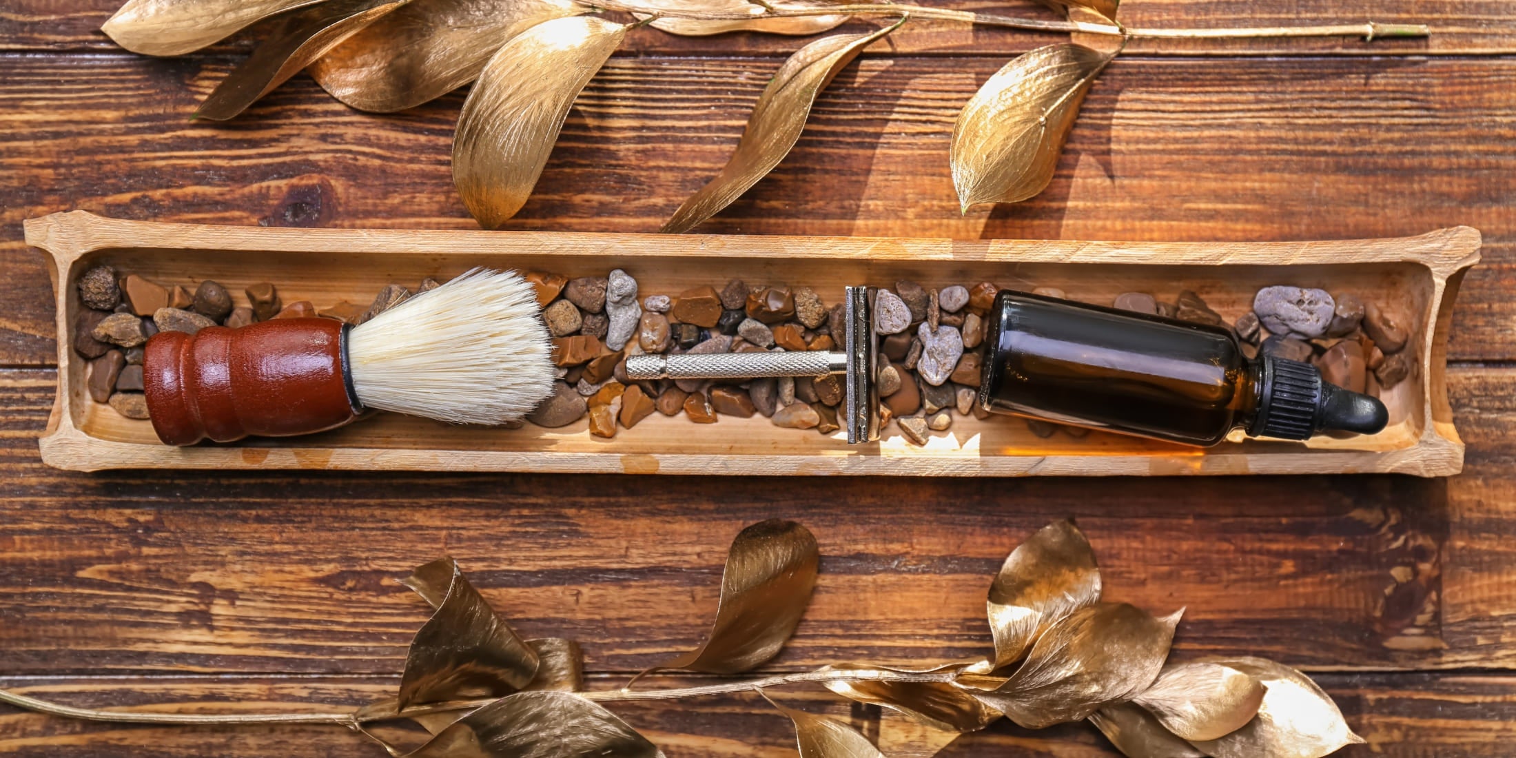 Shaving brush, razor, and a bottle of DIY shave oil neatly arranged on a wooden tray filled with pebbles, surrounded by decorative golden leaves, set on a rustic wooden surface.