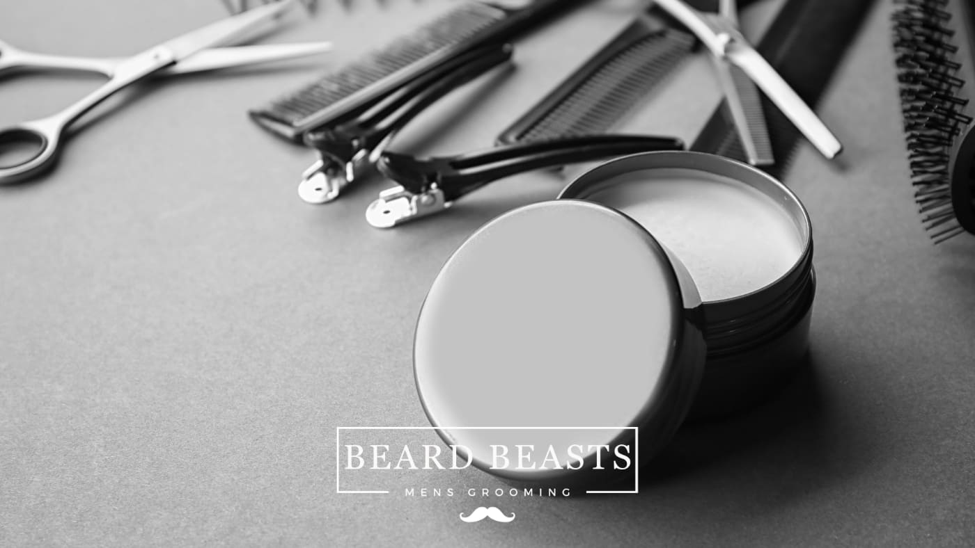 An open tin of pomade next to various hairstyling tools including scissors and combs on a grey background, illustrating the tools and products for men's grooming and how to use pomade.