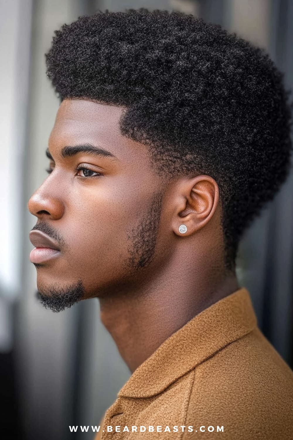 A young man is confidently showcasing a Classic Afro, a timeless and powerful choice among medium length hairstyles for men with naturally curly or coily hair. His Afro is perfectly shaped, highlighting the dense, voluminous texture that makes this style so iconic. The sides are subtly tapered, giving the Afro a neat and defined silhouette that complements his strong facial features. His look is enhanced by a well-groomed beard that mirrors the shape and texture of his hair, creating a harmonious and balanced appearance. He’s wearing a warm, textured jacket that adds to the overall polished yet natural vibe.