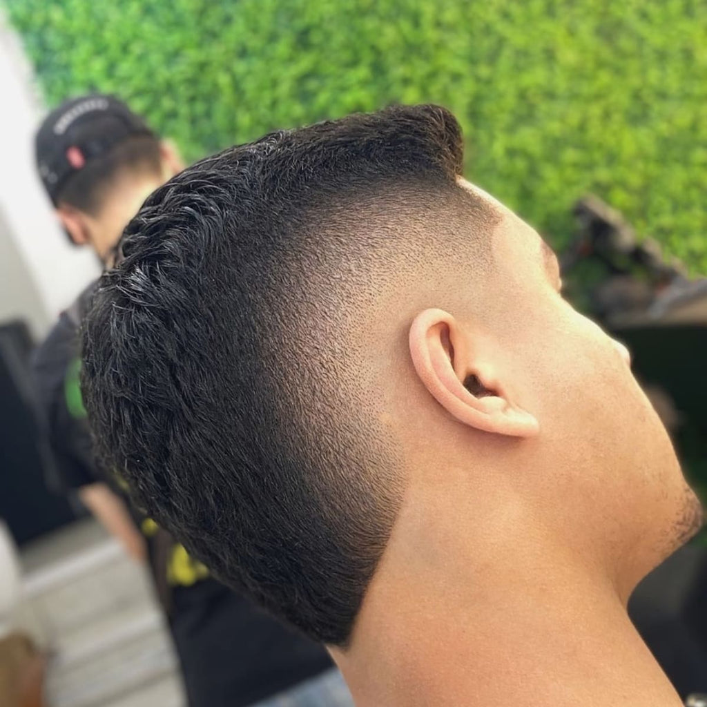 Close-up of a man with a bald burst fade haircut, featuring a rounded fade effect that radiates out around the ear, creating a distinct circular shape. The burst fade contrasts neatly with the textured top, highlighting volume and adding an edgy, modern vibe. Perfect for those wanting a bold, unique hairstyle, this bald burst fade is ideal for drawing attention to the natural contour around the ear.