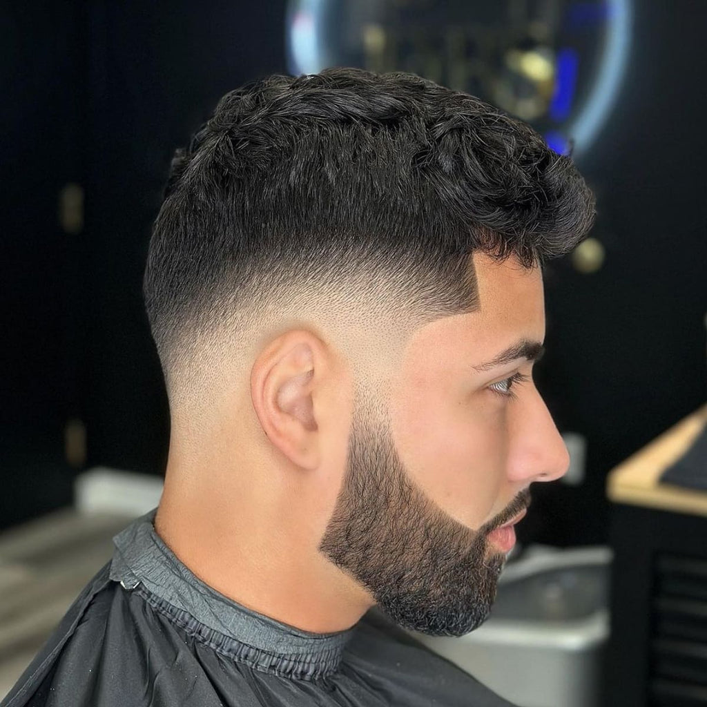 Profile view of a man with a stylish bald drop fade haircut, showcasing a smooth fade that follows the natural curve of the head and dips around the ear for a unique 'drop' effect. The fade transitions seamlessly into a thick, well-groomed beard, emphasizing sharp lines and clean edges. This bald drop fade adds depth and structure, perfect for anyone looking for a modern, polished look that stands out.