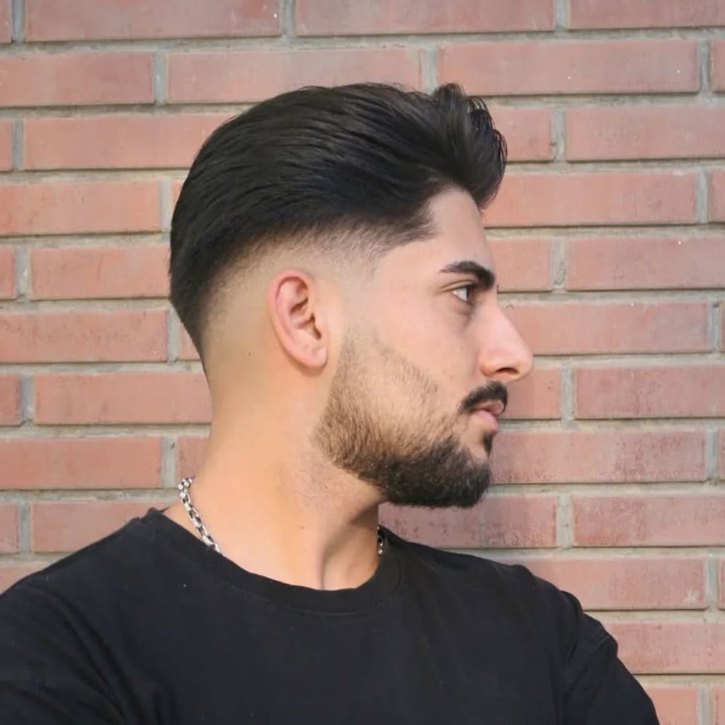 Side profile of a man with a sleek bald drop fade haircut paired with a voluminous pompadour. The clean taper curves smoothly around the sides and back, creating a sharp contrast with the neatly styled, lifted hair on top. The beard is well-groomed and complements the polished, modern look. A timeless and sophisticated style ideal for formal and casual settings alike.