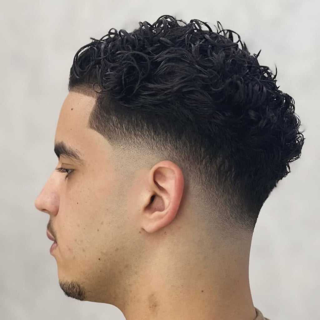 Side profile of a man with a clean and sharp haircut featuring a smooth taper around the sides and back. The top is styled with natural curls, adding volume and texture for a dynamic, stylish look. The lineup at the hairline is precise, complementing the overall polished appearance. This haircut balances natural texture with a neat and modern finish.