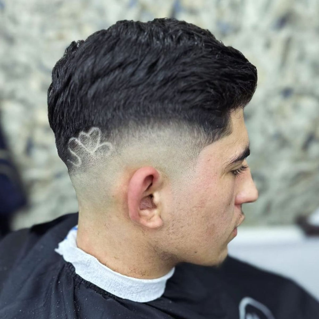 Side profile of a man showcasing a stylish bald drop fade haircut with a unique shaved design on the tapered section. The design features a creative heart-shaped pattern etched into the fade, adding a bold and artistic touch. The clean taper transitions smoothly into the longer hair on top, creating a sharp and polished appearance. A modern and eye-catching style perfect for self-expression.