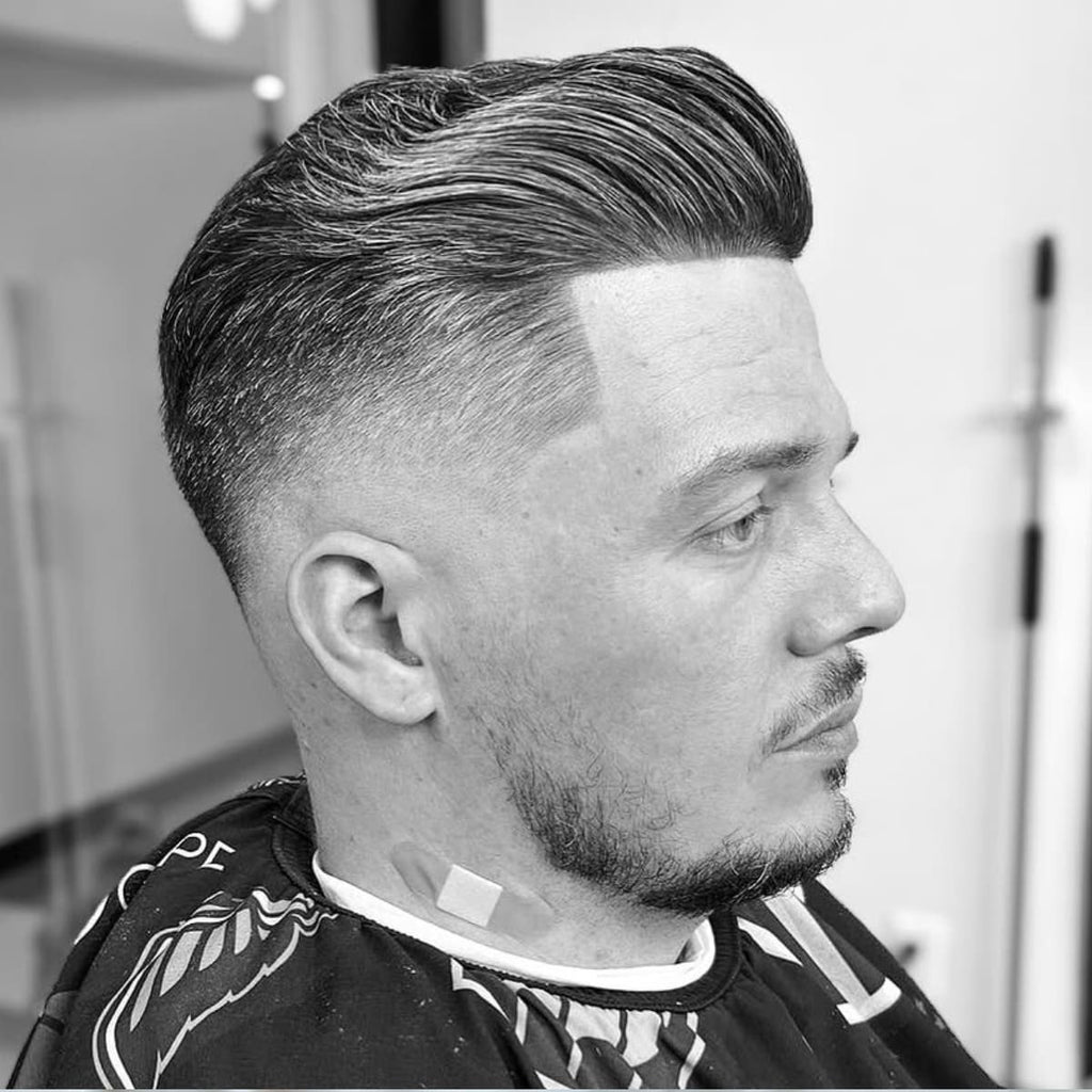 Side profile of a man with a sophisticated bald drop fade paired with slicked-back hair. The haircut features a precise lineup and a smooth fade that curves elegantly around the back of the head. The slicked-back top adds a polished and professional touch, perfect for formal or stylish casual occasions. A timeless look that blends sharp precision with classic elegance.