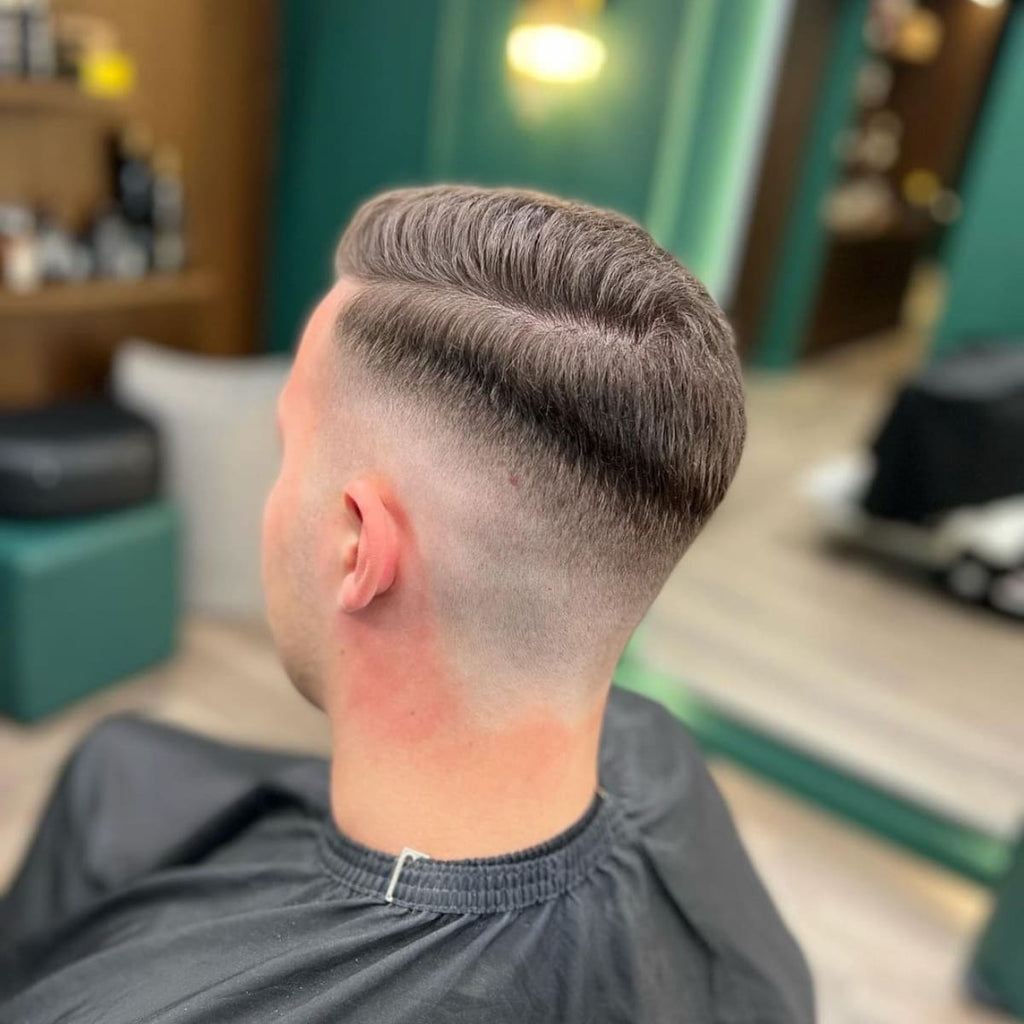 Back view of a man with a neatly styled haircut featuring a hard part and a smooth fade. The haircut shows a clean, sharp parting line that separates the longer, combed-over top from the closely faded sides, creating a polished and structured look. The style emphasizes precision and a professional finish, perfect for a refined yet modern appearance.