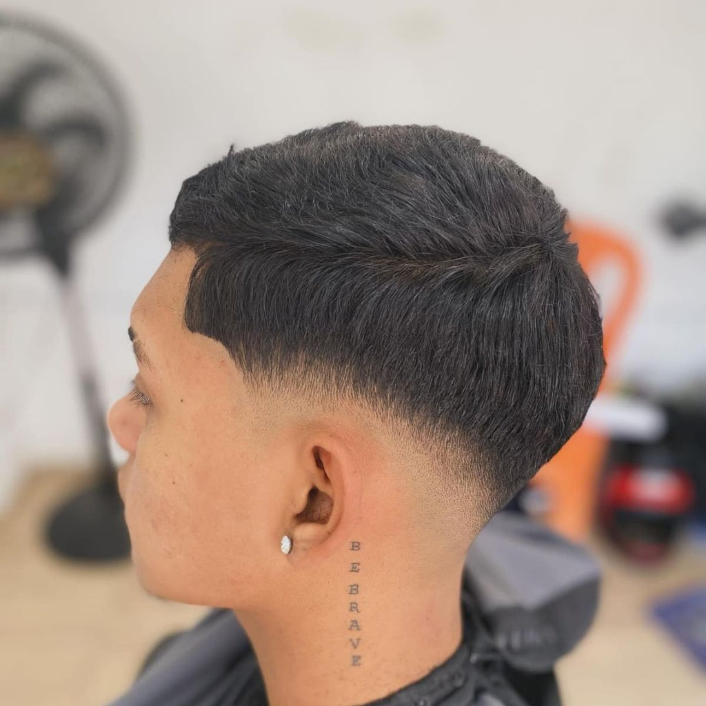 Side profile of a young man with a clean bald low fade haircut, featuring a subtle fade starting near the ears and blending smoothly into the top. The hairstyle highlights a sharp edge along the hairline and a defined part for added structure. Perfect for a sleek, professional look, this low bald fade pairs well with the ‘Be Brave’ tattoo behind the ear, adding personality to the style.