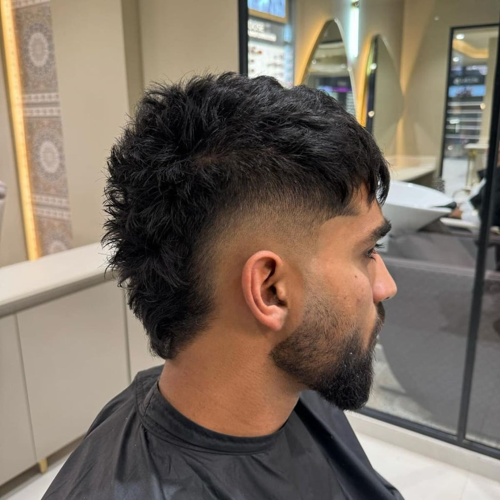 Side profile of a man with a bold bald mohawk fade, featuring a high fade that tapers down to the skin, emphasizing the textured, voluminous mohawk strip along the top. This edgy bald fade haircut accentuates the contrast between the shaved sides and the full, curly mohawk, giving a dynamic and rebellious style. Perfect for those wanting a standout, modern look that combines a mohawk’s attitude with the clean lines of a fade.