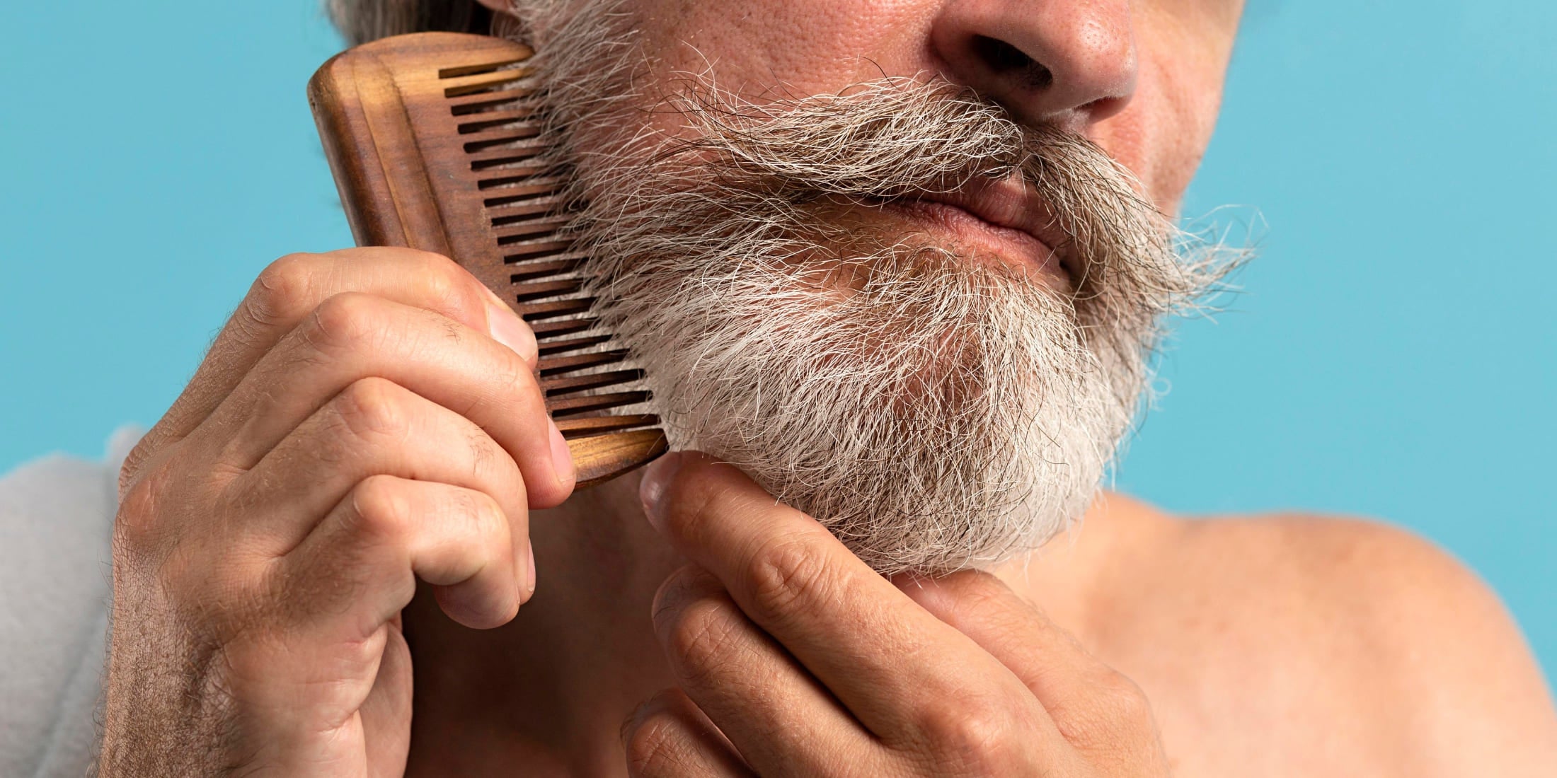 Elderly man with a grey beard combing his beard with a wooden comb, demonstrating proper beard grooming techniques. Learn how to comb your beard effectively for a healthy, well-maintained appearance.