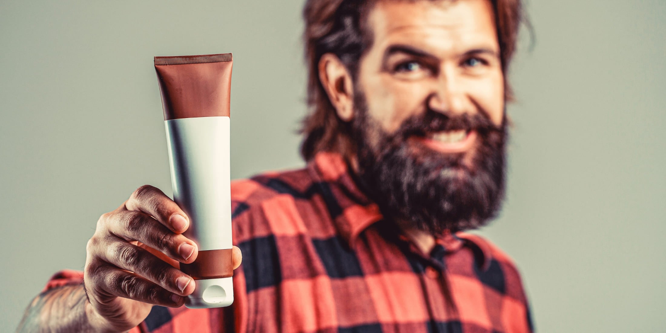 Smiling bearded man in a checkered shirt holding a tube of beard conditioner, illustrating how to use beard conditioner and its benefits for a healthy, soft beard.