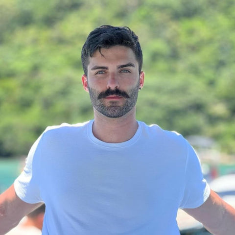 A color portrait of a young man with a stylish beardstache. His beard is short and neatly trimmed, providing a clean look that contrasts with his fuller, prominently styled mustache. The mustache is thick and slightly curled at the ends, adding a classic and sophisticated touch to his appearance. He has dark hair, cut short and styled with a bit of texture. The man is wearing a simple white t-shirt, which enhances his fresh and modern look. He stands against a lush, green, blurred background that suggests an outdoor setting, possibly near a body of water. The lighting is bright and natural, highlighting his facial features and the meticulous grooming of his beard and mustache. His expression is confident and relaxed, with his arms casually resting on a surface, giving off a laid-back yet refined vibe.