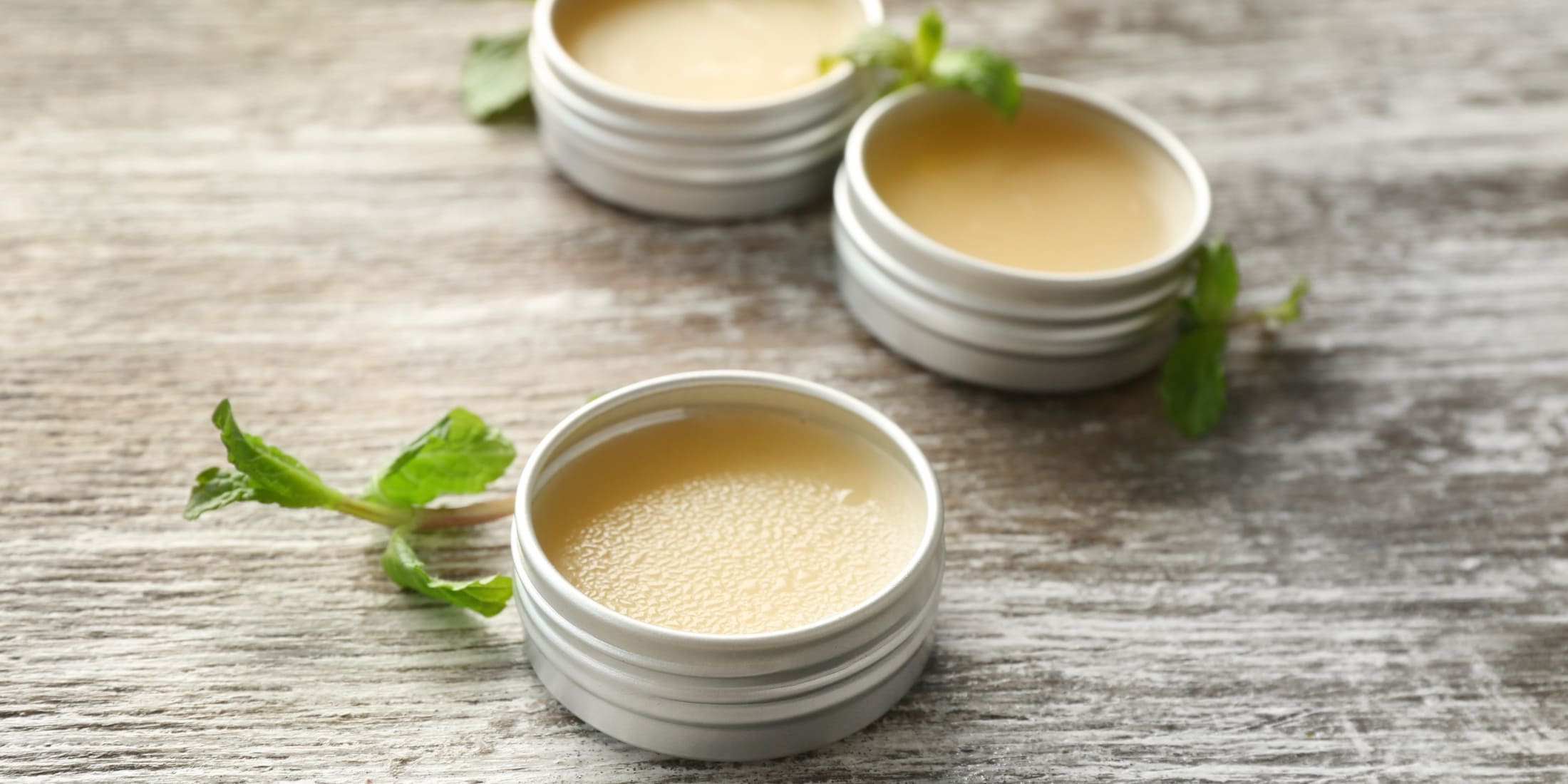 Three small tins of beard balm, placed on a rustic wooden surface, with fresh green mint leaves nearby. The balm in each tin appears smooth and slightly glossy, highlighting its moisturizing properties. Perfect for understanding the benefits in the beard balm vs beard conditioner debate.