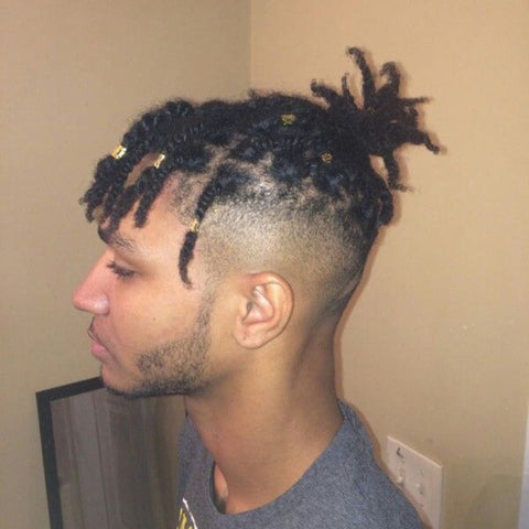 A young man is shown with a braided fringe hairstyle that features short dreadlocks styled into individual braids. The braids are adorned with small, gold hair cuffs, adding a touch of style and personality to the look. His hair is gathered into a small bun at the back, leaving some of the braided fringes to fall naturally over his forehead. The sides of his head are faded close to the skin, creating a clean and sharp contrast between the braids on top and the closely cropped sides. He has a neatly trimmed beard that follows the contours of his jawline, enhancing his well-groomed appearance. The background is a simple, neutral-toned room, which allows the details of his hairstyle to stand out prominently. His expression is calm and focused, perfectly complementing the unique and stylish elements of his braided fringe.