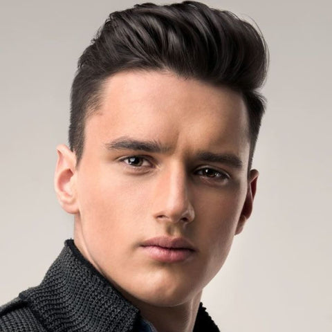 A young man is shown with a brushed-up fringe hairstyle, where the front of his hair is styled upward and back, creating height and volume. His dark hair is meticulously groomed, with the sides tapered and neatly blended into the longer top. The brushed-up fringe gives his look a polished and sophisticated appearance, emphasizing his strong facial features and clean-cut style. He is dressed in a textured, dark knit jacket, which adds a subtle layer of refinement to his overall look. The background is a soft, neutral tone that keeps the focus on his well-styled hair and confident expression. This hairstyle is ideal for men seeking a modern, sleek look that is both versatile and easy to maintain.