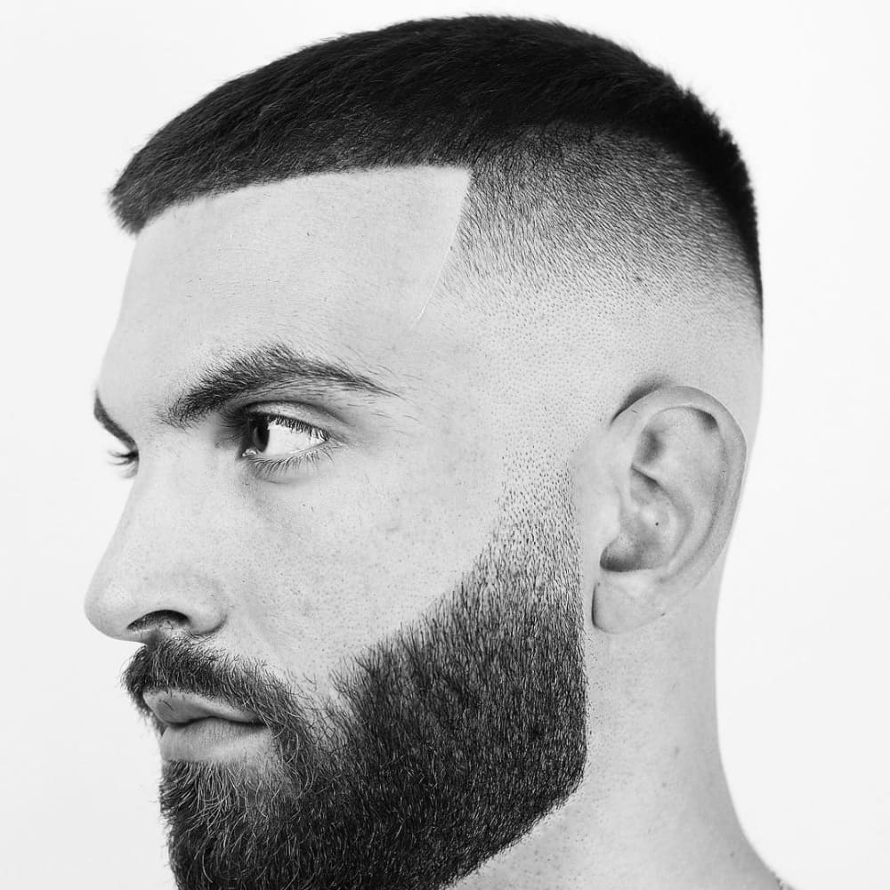 A black-and-white close-up side profile of a man with a butch cut haircut. The haircut features a short, even length on top, transitioning into a precise skin fade on the sides and back. The hairline is sharply defined with a clean line-up at the forehead, creating a crisp, geometric shape. The man also sports a full, neatly trimmed beard that enhances the structured look of the haircut. The overall style is sharp, bold, and modern, emphasizing the contrast between the tightly faded sides and the slightly longer hair on top.