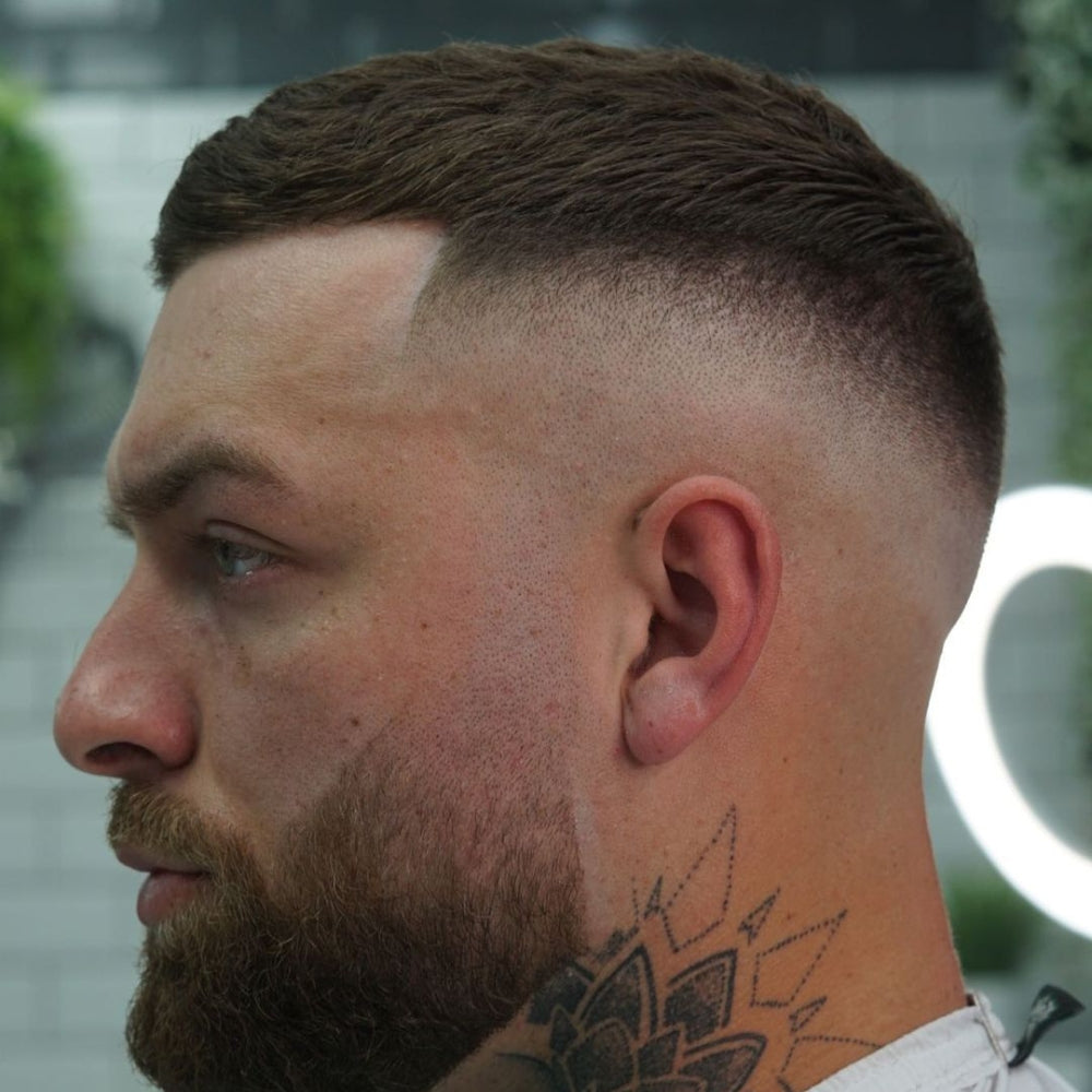 A close-up side profile of a man with a buzz cut featuring a drop fade. The haircut is short and textured on top, with the sides and back gradually fading down to the skin in a smooth drop fade that curves around the ears and nape of the neck. The hairline is sharply defined at the forehead, giving the style a clean and polished appearance.