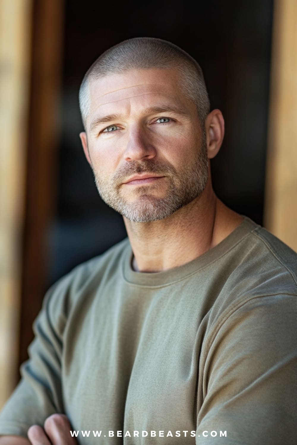 A close-up image of a man with a Buzz Cut hairstyle, showcasing a very short, evenly trimmed haircut that is a classic example of military haircuts for men. The man has a well-groomed, light beard that enhances his strong jawline, further emphasizing the sharp and clean look associated with this style. He is dressed in a simple olive-green t-shirt, which complements his rugged, yet polished appearance. The background is softly blurred, ensuring the focus remains on his Buzz Cut and overall masculine, no-nonsense look, perfectly illustrating the practicality and timeless appeal of short military haircuts for men.