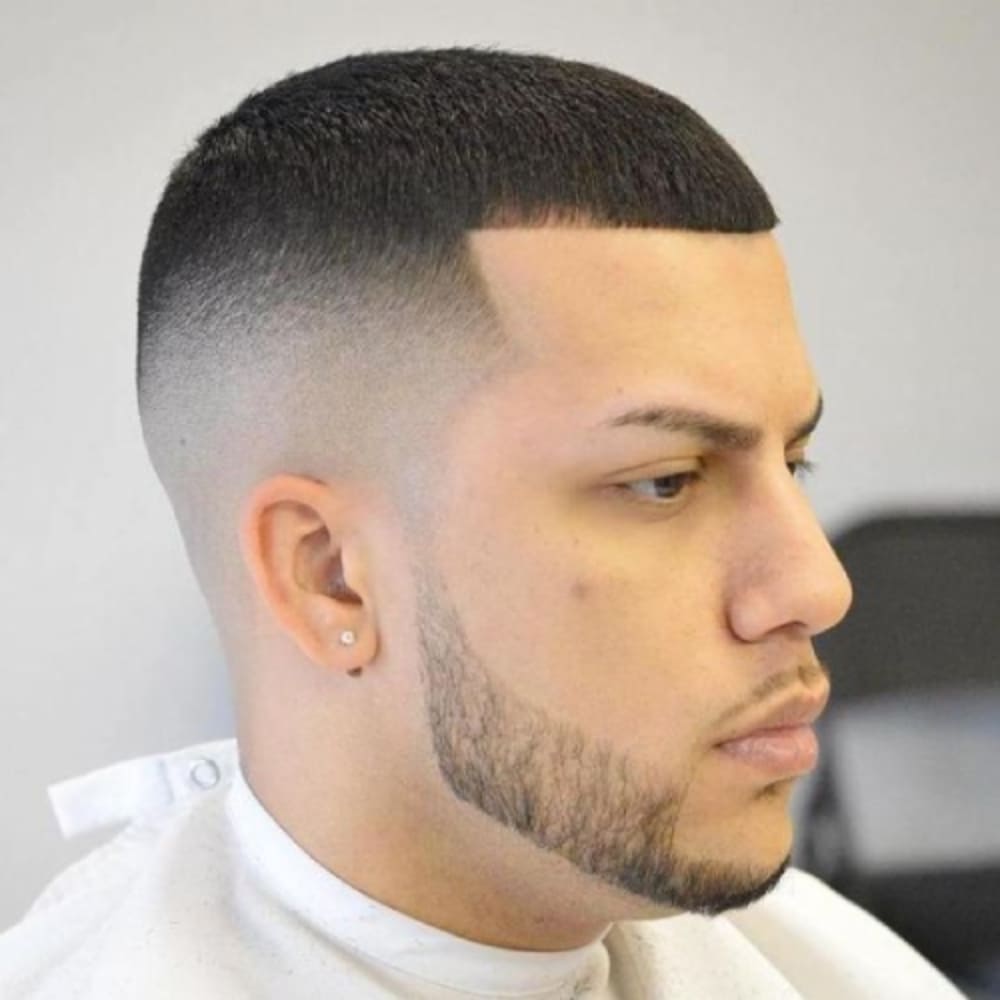 A close-up side profile of a man with a buzz cut featuring a precise line-up and high fade. The haircut is short and uniform on top, with a sharply defined, straight line-up at the forehead and temples that gives the style a clean, angular appearance. The sides and back are blended into a high skin fade, creating a smooth transition from the top to the fade.