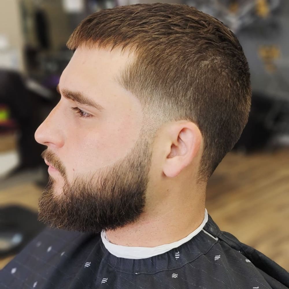 A close-up side profile of a man with a buzz cut featuring a temple fade. The haircut has a short, even length on top with a subtle fade starting at the temples and blending smoothly into the sides and back. The temple fade is clean and precise, adding a modern touch to the classic buzz cut.