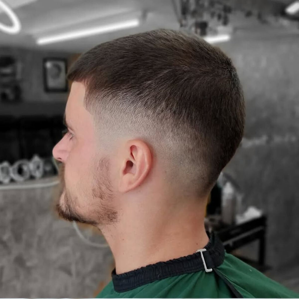 Side profile of a man with a clean and modern bald drop fade haircut paired with a short buzz cut. The haircut features a smooth transition from the slightly longer hair on top to a precise skin fade that curves naturally around the back of the head. The style emphasizes sharp lines and a neat, polished appearance. A low-maintenance yet stylish look ideal for a fresh and professional vibe.