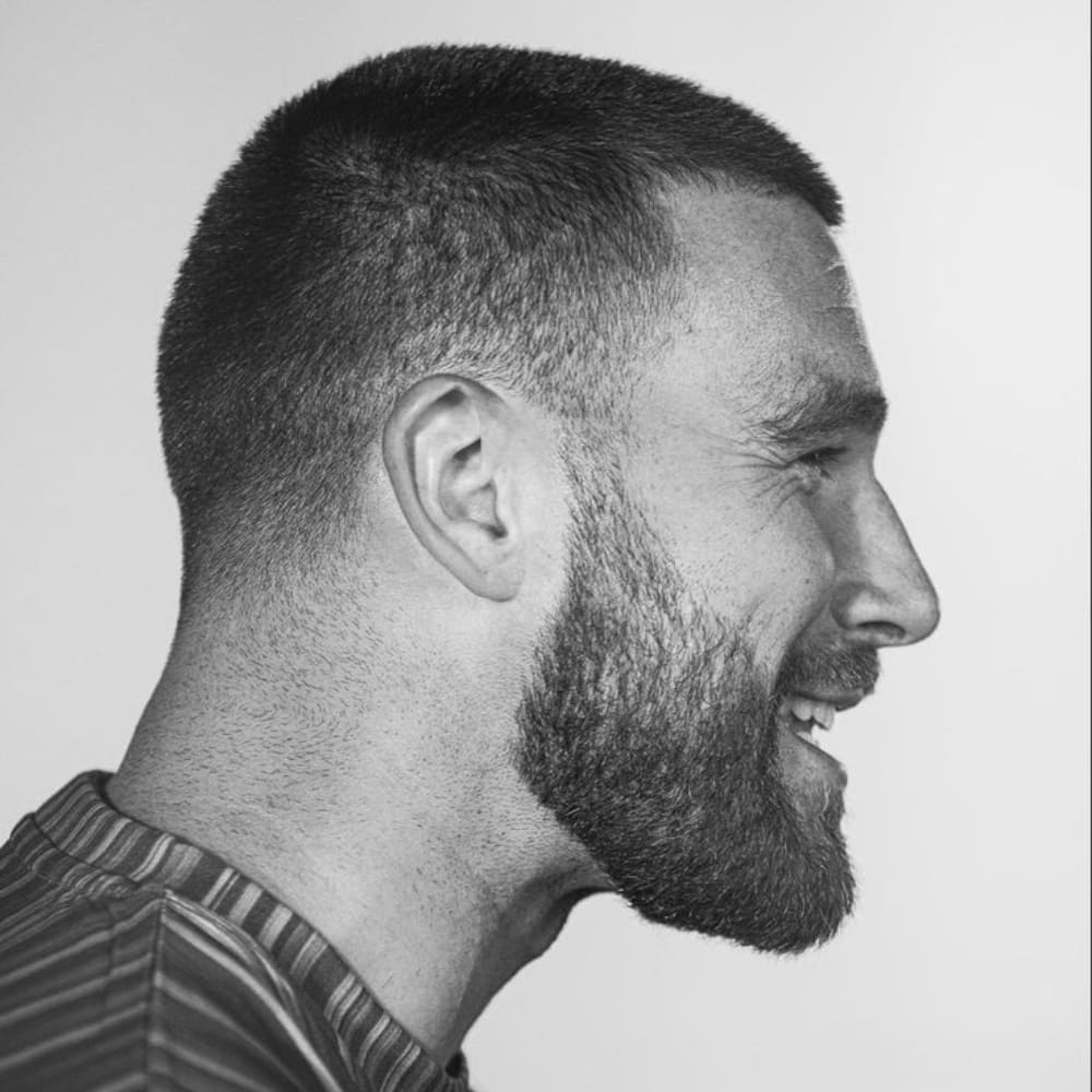 A black-and-white side profile of a man with a buzz cut paired with a full beard. The buzz cut is neatly trimmed, short, and uniform, giving a clean and minimalist appearance. The man's beard is thick, well-groomed, and complements the short hair, adding a rugged and masculine contrast. He is smiling, which highlights the sharpness of the beard line and the tidy cut of the hair. The overall look is stylish, modern, and exudes confidence, with the simplicity of the buzz cut balanced by the fullness of the beard.