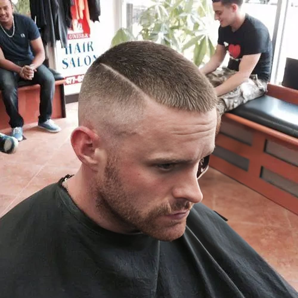 A close-up image of a man with a buzz cut with a hard part. The haircut features short, even hair on top with a clean, sharp line shaved into the side of the head, creating a distinct hard part. The sides and back are blended into a high fade, adding to the modern and stylish look. The man's facial hair is trimmed to a short, stubbly beard, complementing the neat and polished haircut. The hard part adds a unique and edgy twist to the classic buzz cut, making this style both bold and fashionable. The setting is a barbershop with other patrons in the background.