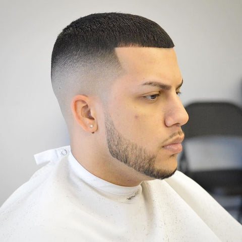 Man with a Caesar haircut featuring a very short, straight fringe and a sharp high fade on the sides and back. The hair on top is cut close to the scalp, resembling a buzz cut. He has a neatly trimmed beard and a small stud earring, creating a clean and polished look. The background is simple, focusing attention on the precise haircut