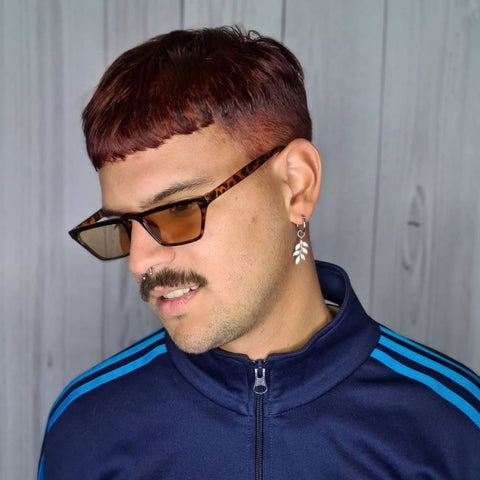 A man is wearing a Caesar cut, a fringe haircut characterized by a short, straight-across fringe that sits just above the forehead. His hair is a deep reddish-brown color, and the sides are trimmed short, contrasting with the slightly longer hair on top. He is accessorized with angular tortoiseshell sunglasses and a dangling earring shaped like a leaf on his left ear. He also sports a neat mustache and a small septum ring. He is dressed in a navy blue track jacket with light blue stripes on the shoulders, standing against a light wood-paneled background.