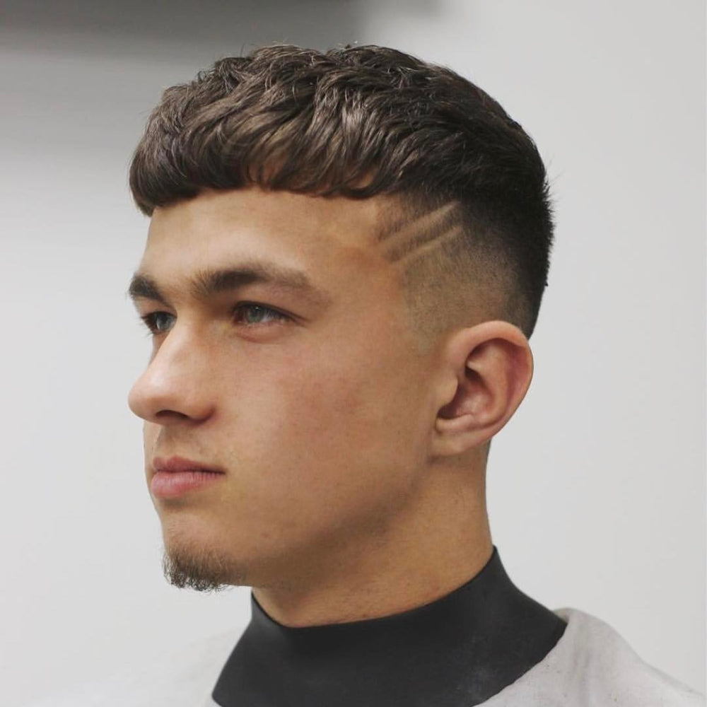 Young man with a Caesar crop haircut, featuring a textured top with choppy layers and a straight fringe. The sides are faded with two shaved lines as a design element, creating a modern and stylish look