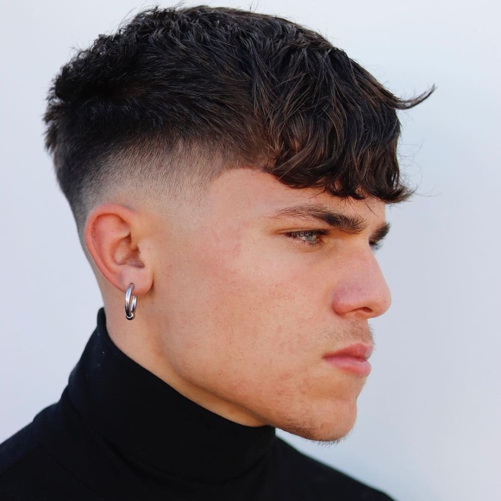 Young man with a Caesar haircut featuring a textured, wavy top and a drop fade on the sides and back. The fringe is styled with natural waves, adding volume and movement. He is wearing a black turtleneck and a single silver hoop earring, standing against a plain white background, which highlights the clean lines and stylish contrast of his haircut.