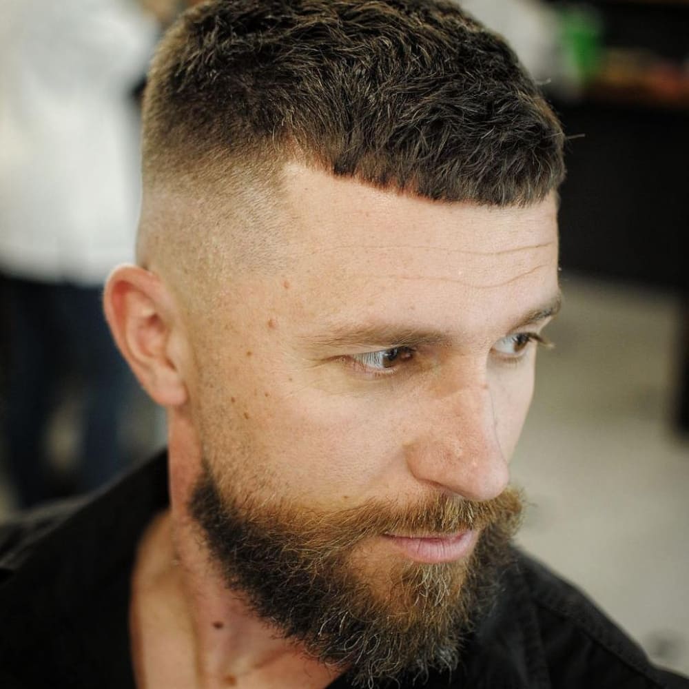 Man with a Caesar haircut featuring a short, straight fringe and a high fade on the sides and back. He has a full, well-groomed beard that complements the clean lines of the haircut, creating a rugged yet polished look.