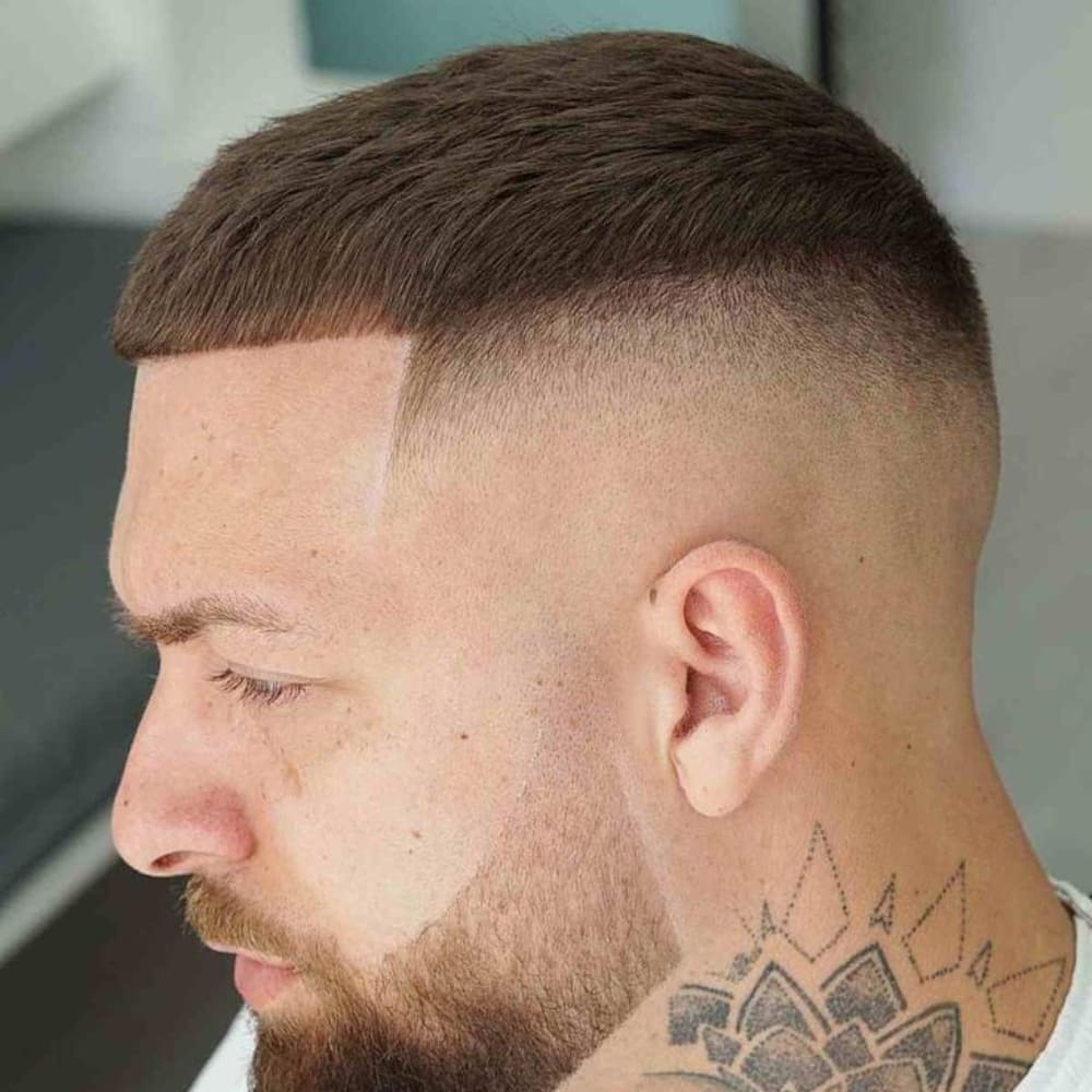 Man with a Caesar haircut featuring a short, straight fringe and a high fade on the sides and back. The haircut is complemented by a neatly trimmed beard and a geometric neck tattoo, creating a modern and stylish look.